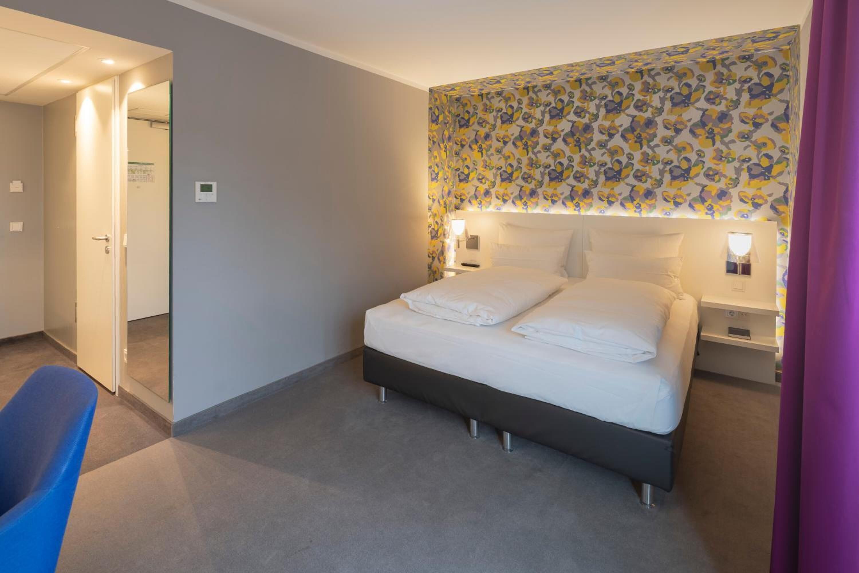 A hotel room with a bed, bedside tables, a chair, and a patterned wall.