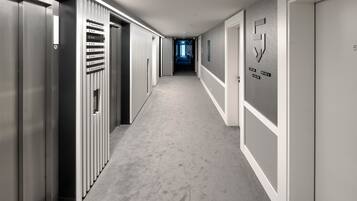 Un couloir d’hôtel moderne, avec un long couloir, des portes d’ascenseur et un panneau mural indiquant les numéros de chambre.