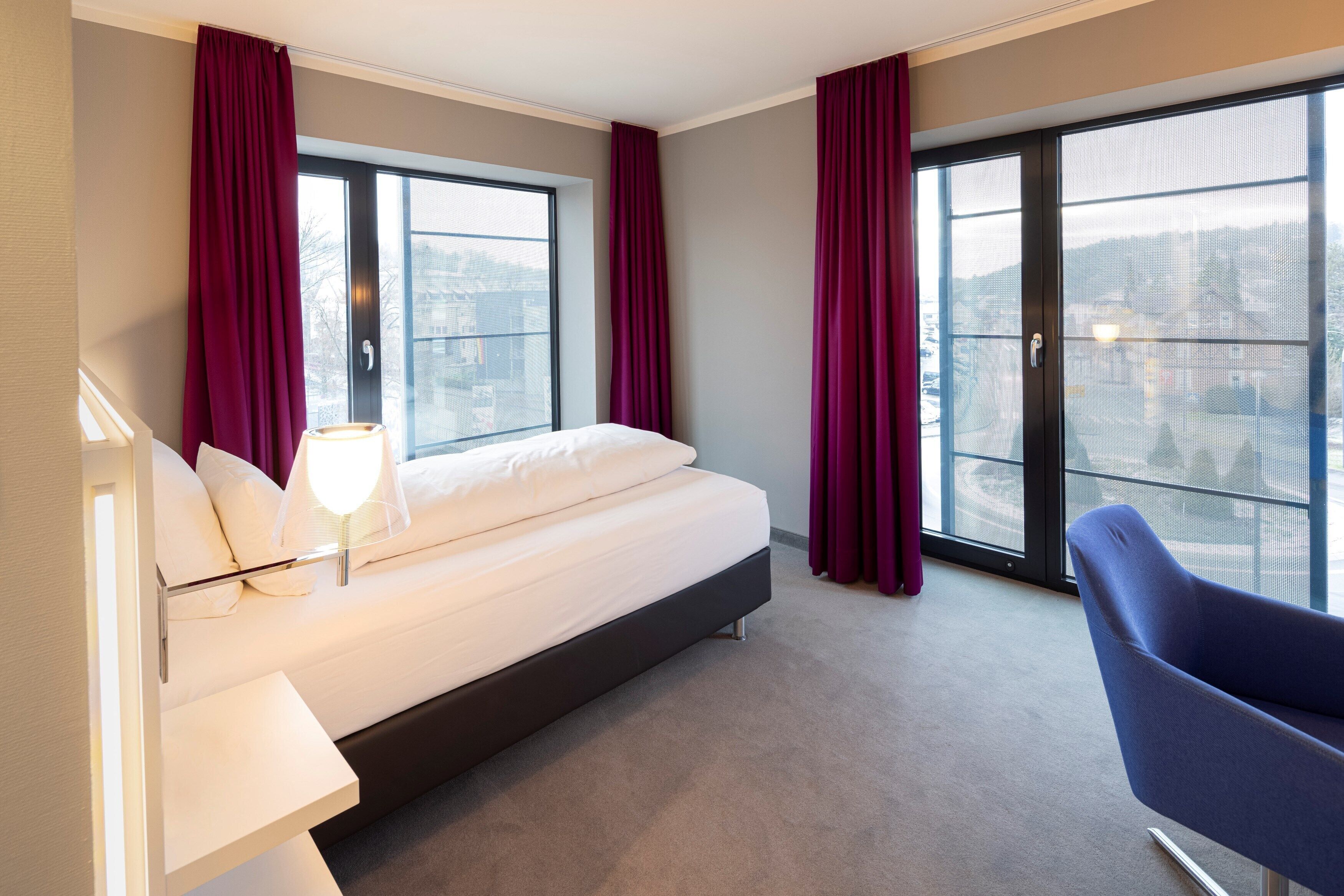 A modern hotel room with a large bed, a chair, a desk, and a view of the city through large windows.