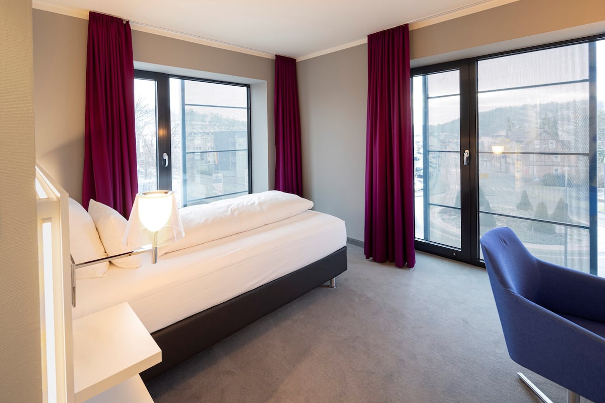 Ett modernt hotellrum med en stor säng, en stol, ett skrivbord och utsikt över staden genom stora fönster.