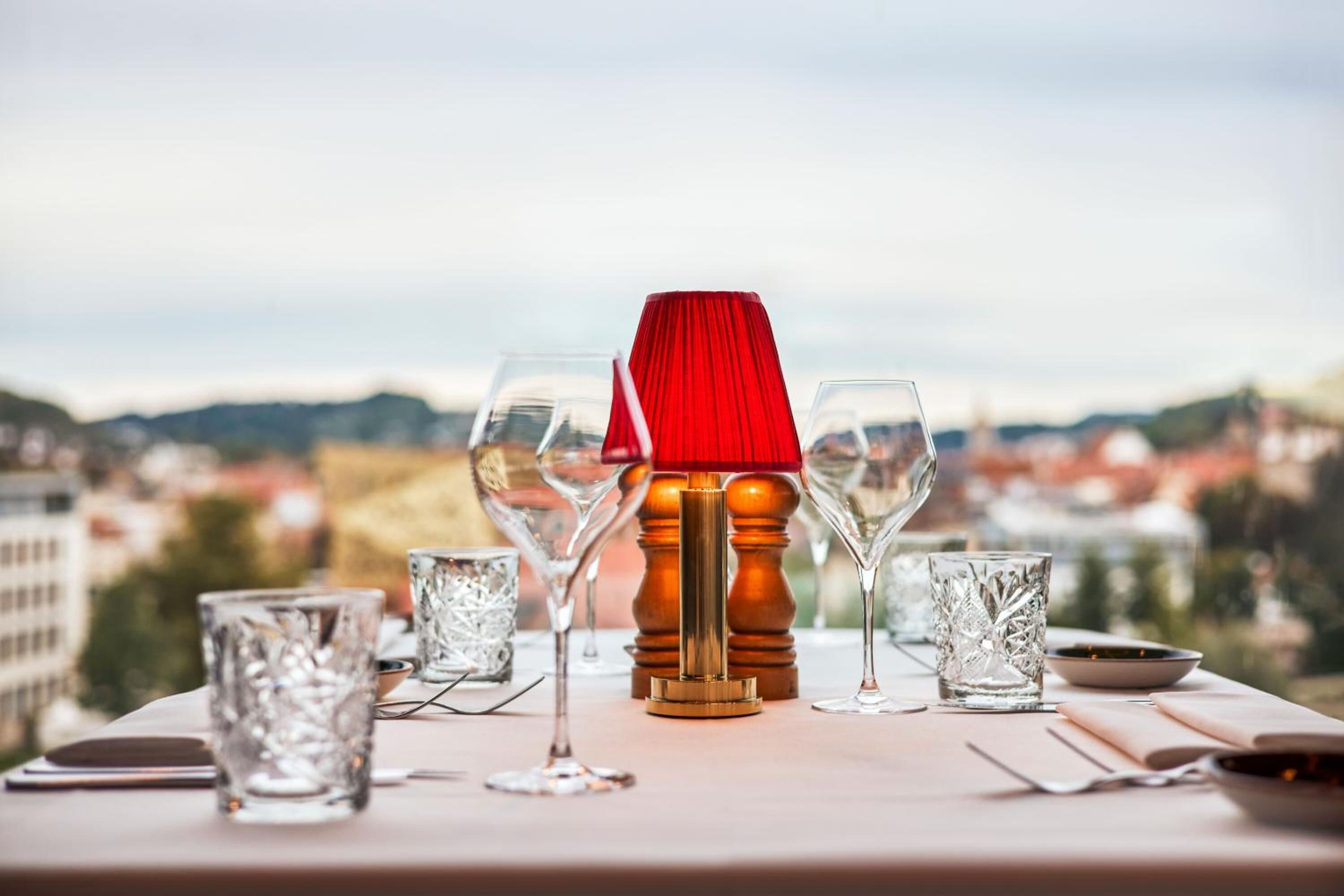 Een tafel gedekt met wijnglazen en een rode lamp, met uitzicht op een stadsbeeld.