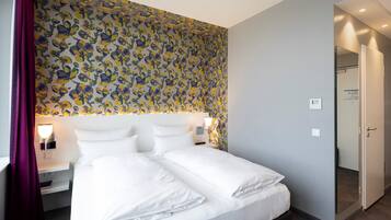 A hotel room with a large bed, a headboard with a patterned wallpaper, bedside lamps, and a door to the right.