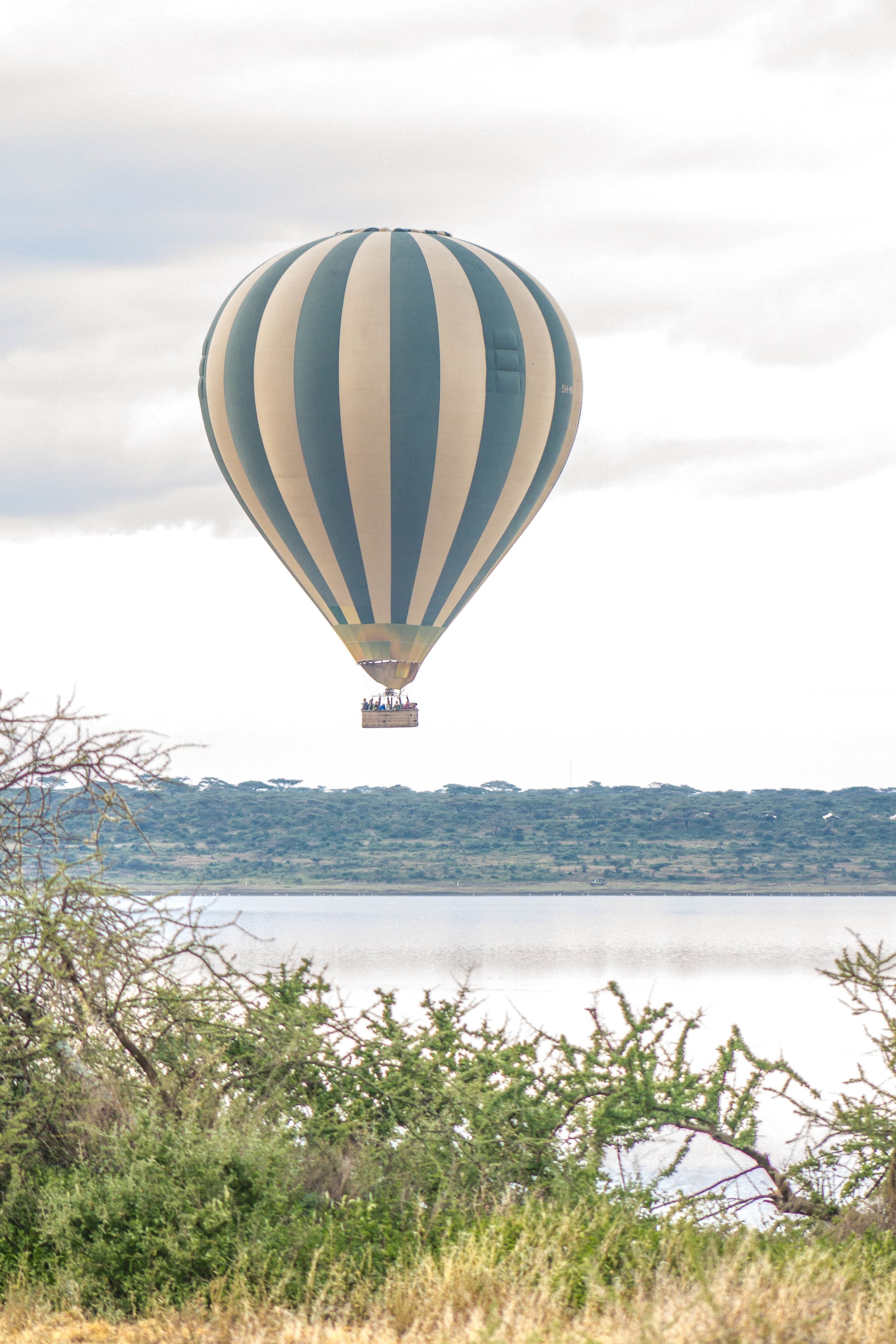 A hot air balloon with a striped pattern flying over a body of water, with trees and a distant landscape in the background.