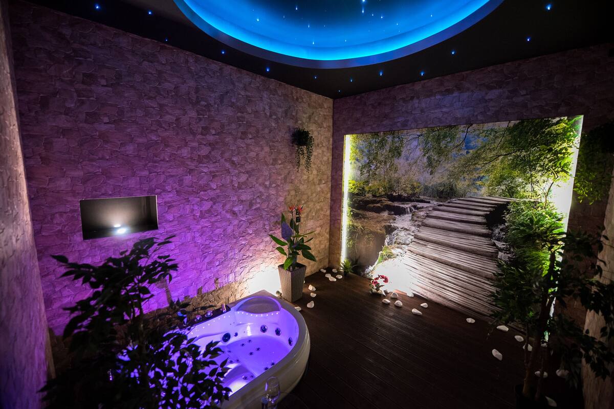 A spa room with a stone wall, a large window with a waterfall view, a hot tub, and a ceiling with star-like lights.