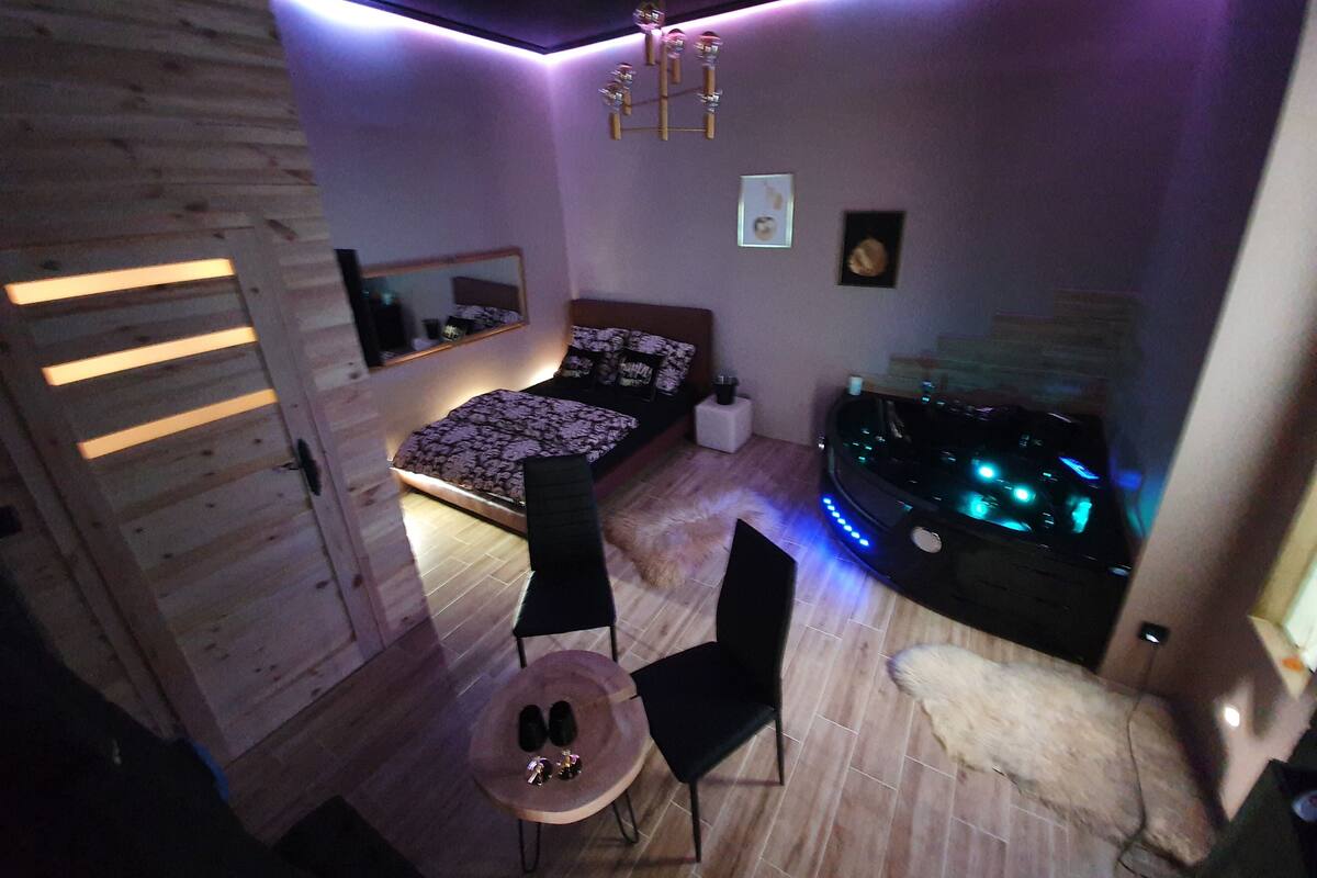 A bedroom with a bed, a chair, a small table, a vanity with a mirror, and a wall-mounted heater.