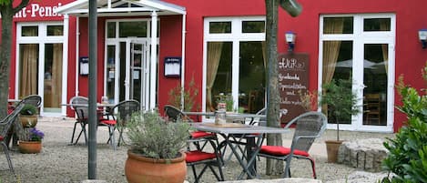 An outdoor seating area with tables and chairs, potted plants, and a red building facade.