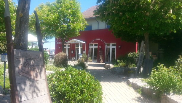 A red building with a white entrance, surrounded by greenery and a wooden sign with a horse. - Pension A'MiRo (Rust)