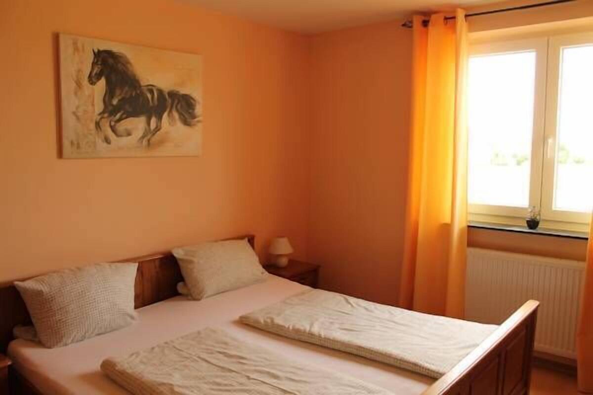 A bedroom with a bed, pillows, a nightstand, a lamp, a window with curtains, and a painting on the wall.