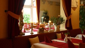 A dining area with red curtains, a vase with a plant, candles, and a statue on the table. - Pension A'MiRo (Rust)