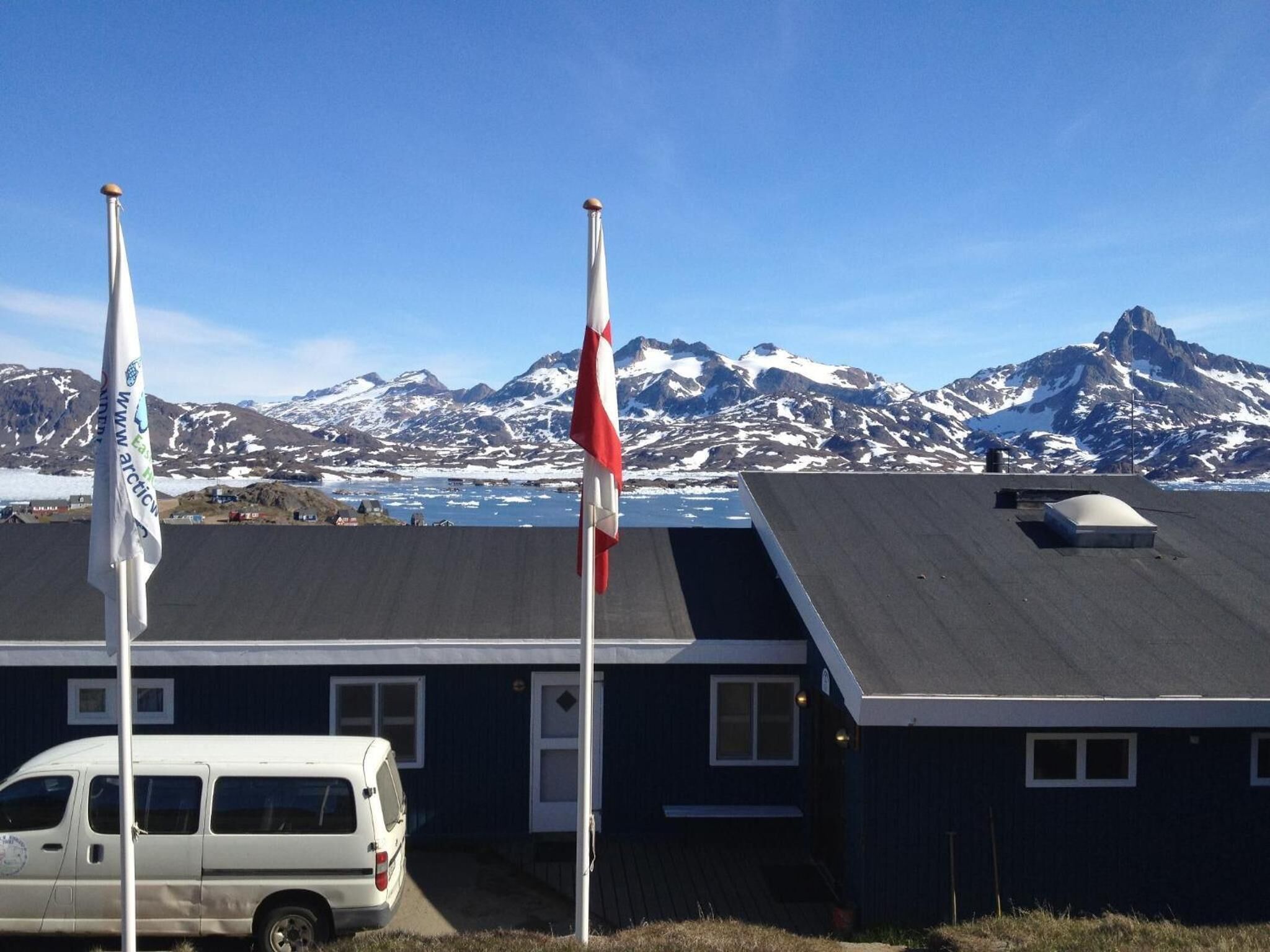 A building with a dark exterior and a sloped roof, a white van parked in front, and two flags on flagpoles.
