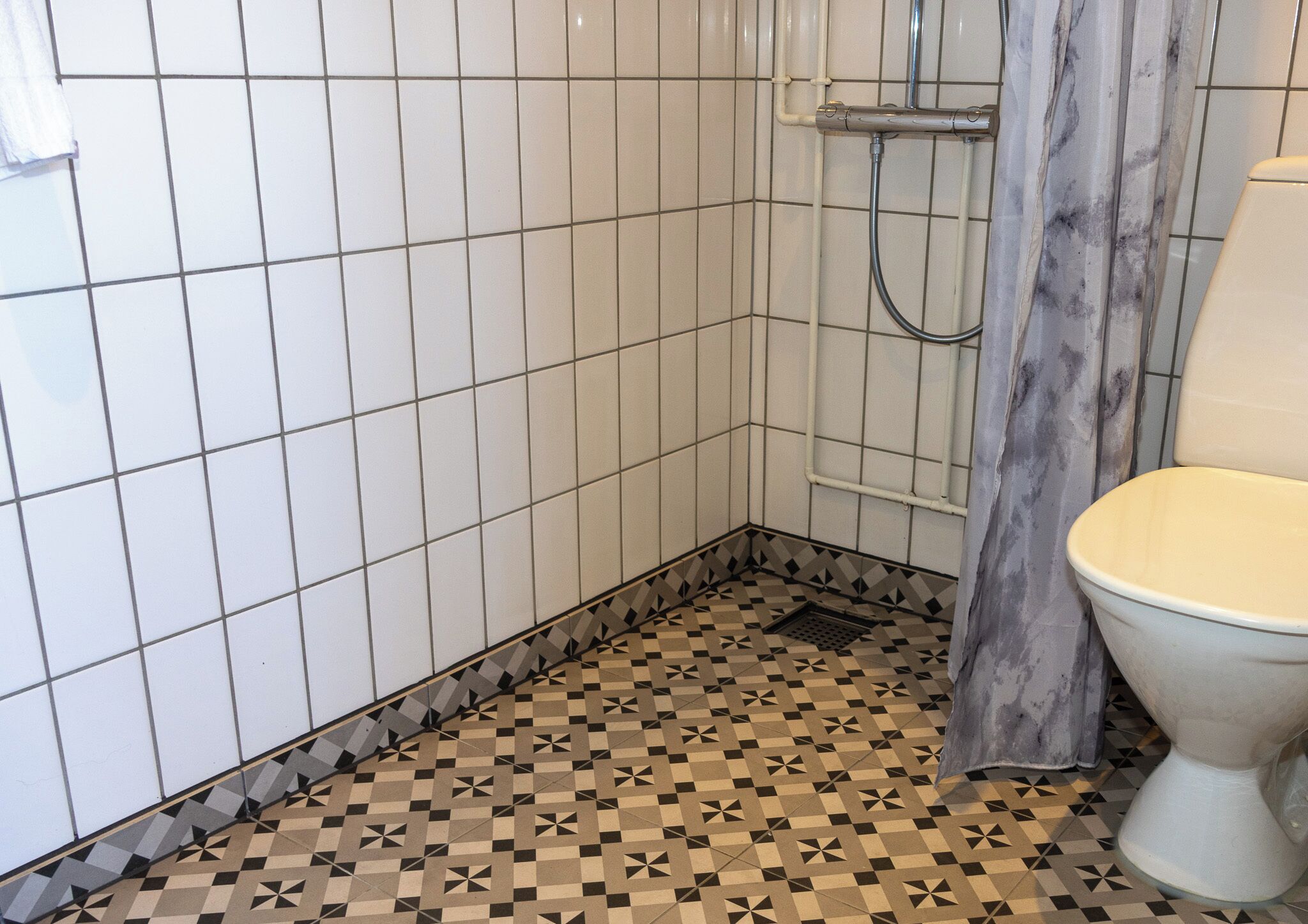 A bathroom with white tiled walls, a patterned floor, a shower with a curtain, and a toilet.