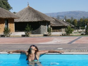 Uma piscina com uma pessoa aproveitando a água, cercada por cabanas com telhado de palha e uma paisagem montanhosa ao fundo.