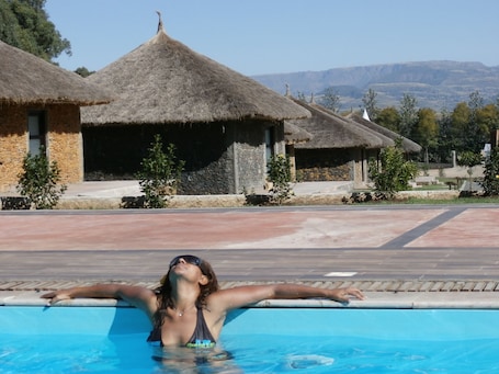 A swimming pool with a person enjoying the water, surrounded by thatched-roof huts and a mountainous landscape in the background.