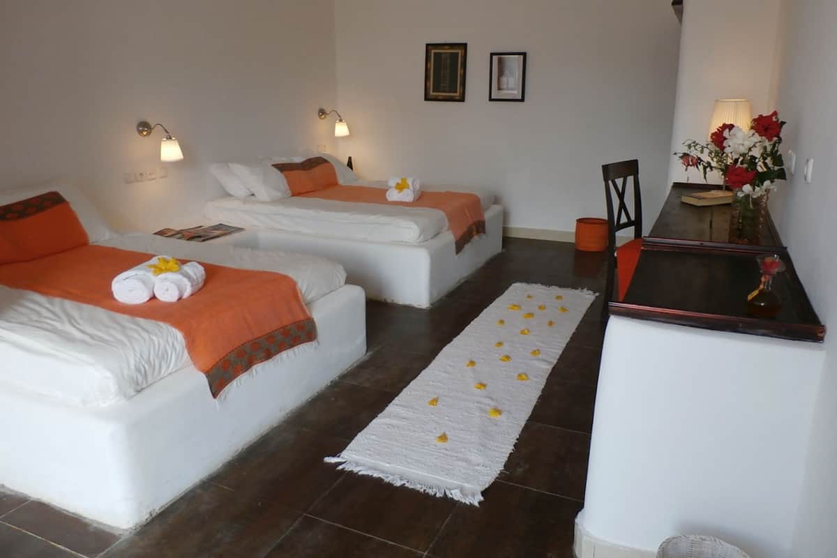 Habitación de hotel con dos camas, una mesita pequeña y una silla. Hay luces fijadas a la pared y un florero con flores sobre la mesita.