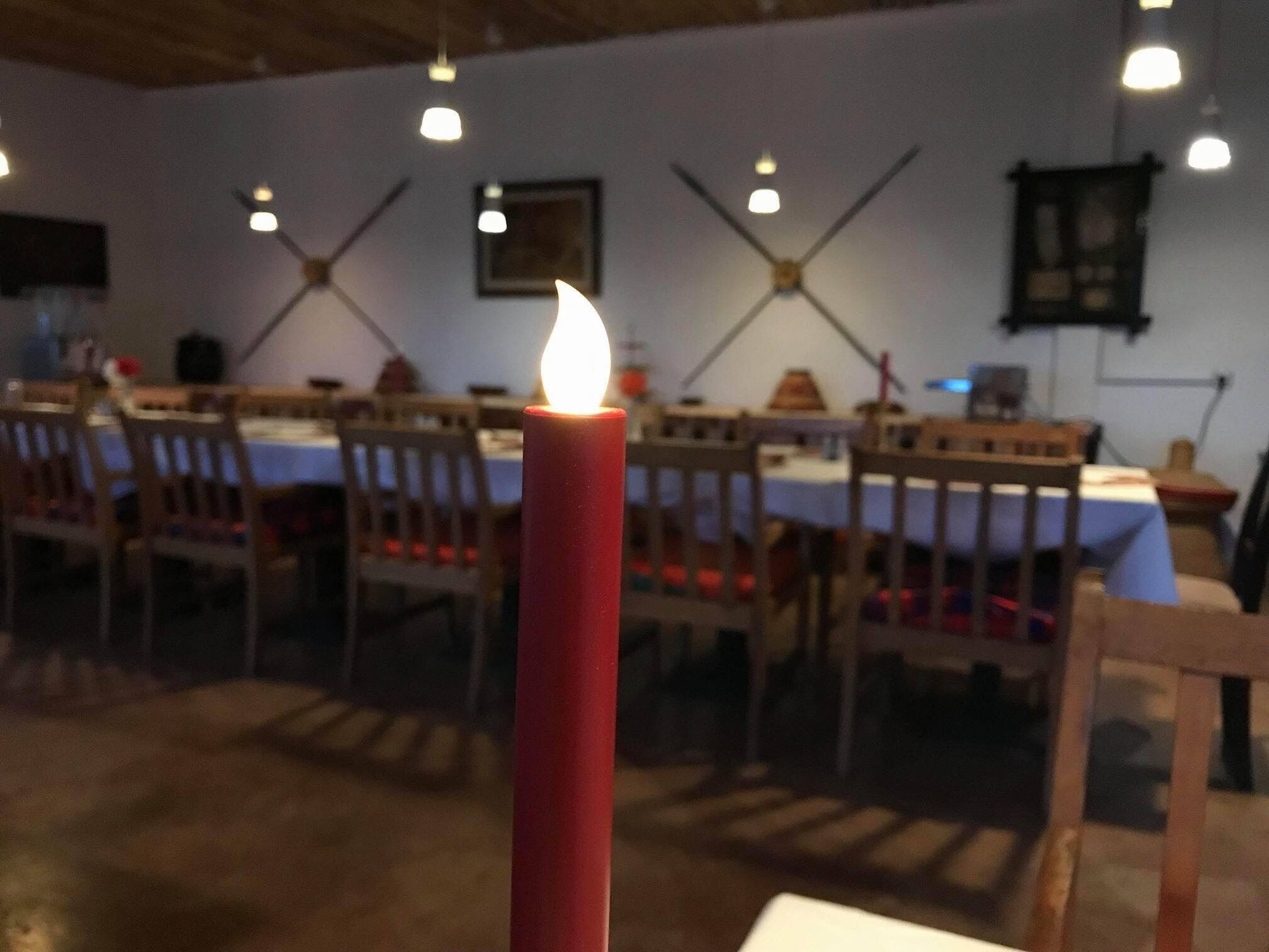 Eine Kerze auf einem Tisch in einem Raum mit Holztischen und -stühlen, einem Deckenventilator und Pendelleuchten.