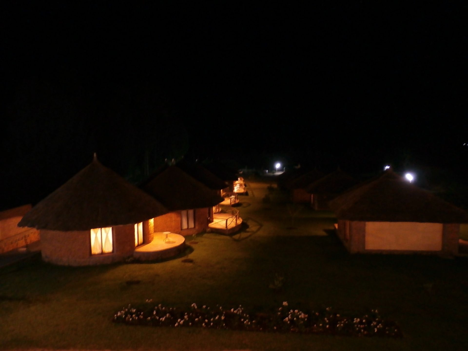 A row of thatched-roof huts with illuminated windows, arranged in a linear formation on a grassy area at night.