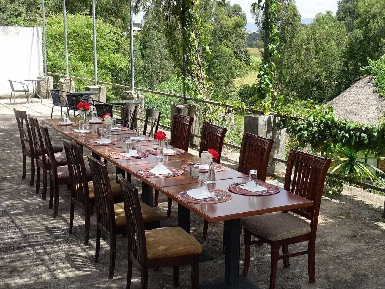 An outdoor dining area with a long wooden table set for a meal, surrounded by chairs.
