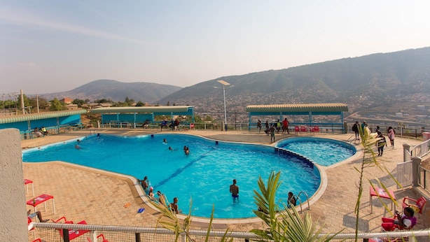 A swimming pool with multiple lanes, surrounded by red chairs and umbrellas, with a view of distant mountains.