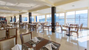 A spacious dining area with multiple tables and chairs, large windows offering a view of the sea and mountains, and a clean, modern interior design.