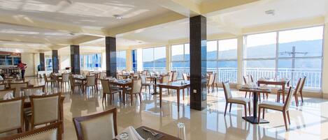Un vaste espace dédié à la salle à manger, avec de nombreuses tables et chaises, de grandes fenêtres offrant une vue sur la mer et les montagnes, et un aménagement intérieur propre et moderne.
