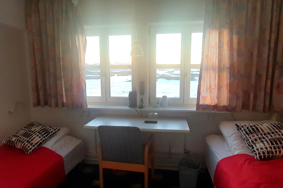 Een kamer met twee bedden, een bureau en een stoel. Er is een raam met gordijnen en uitzicht op zee.