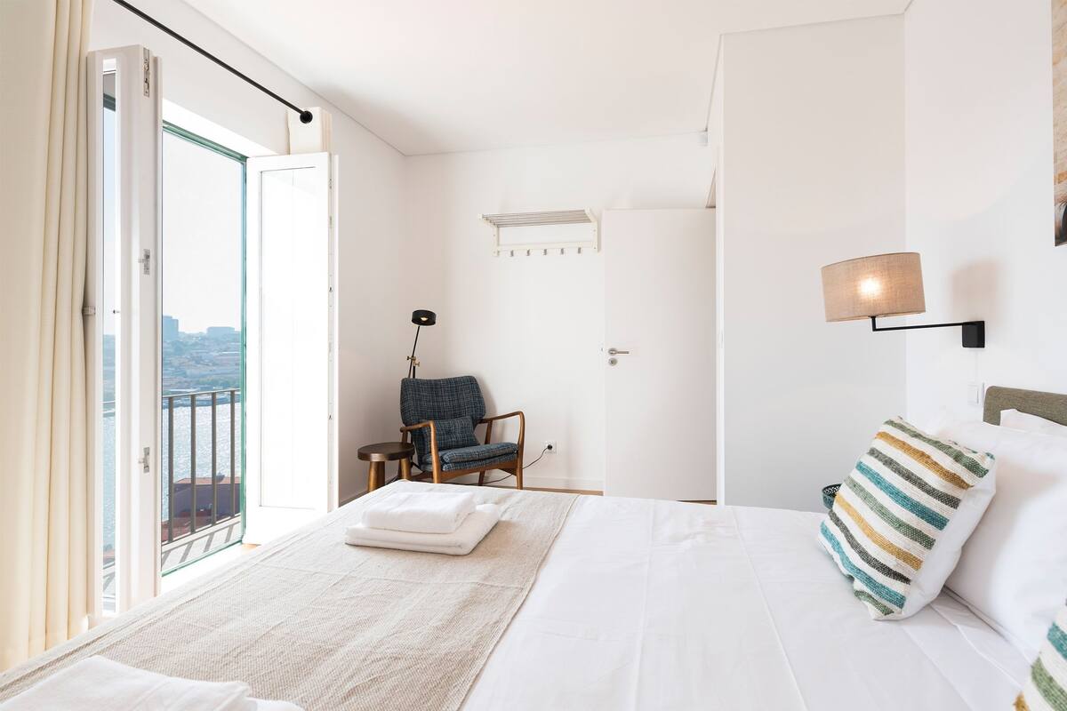 A hotel room with a bed, a chair, a lamp, and a view of the city through a large window.