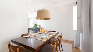 A dining area with a wooden table set for four, a large hanging lamp, and a view of a living room with a sofa and a plant. - Arco Apartments (Porto)