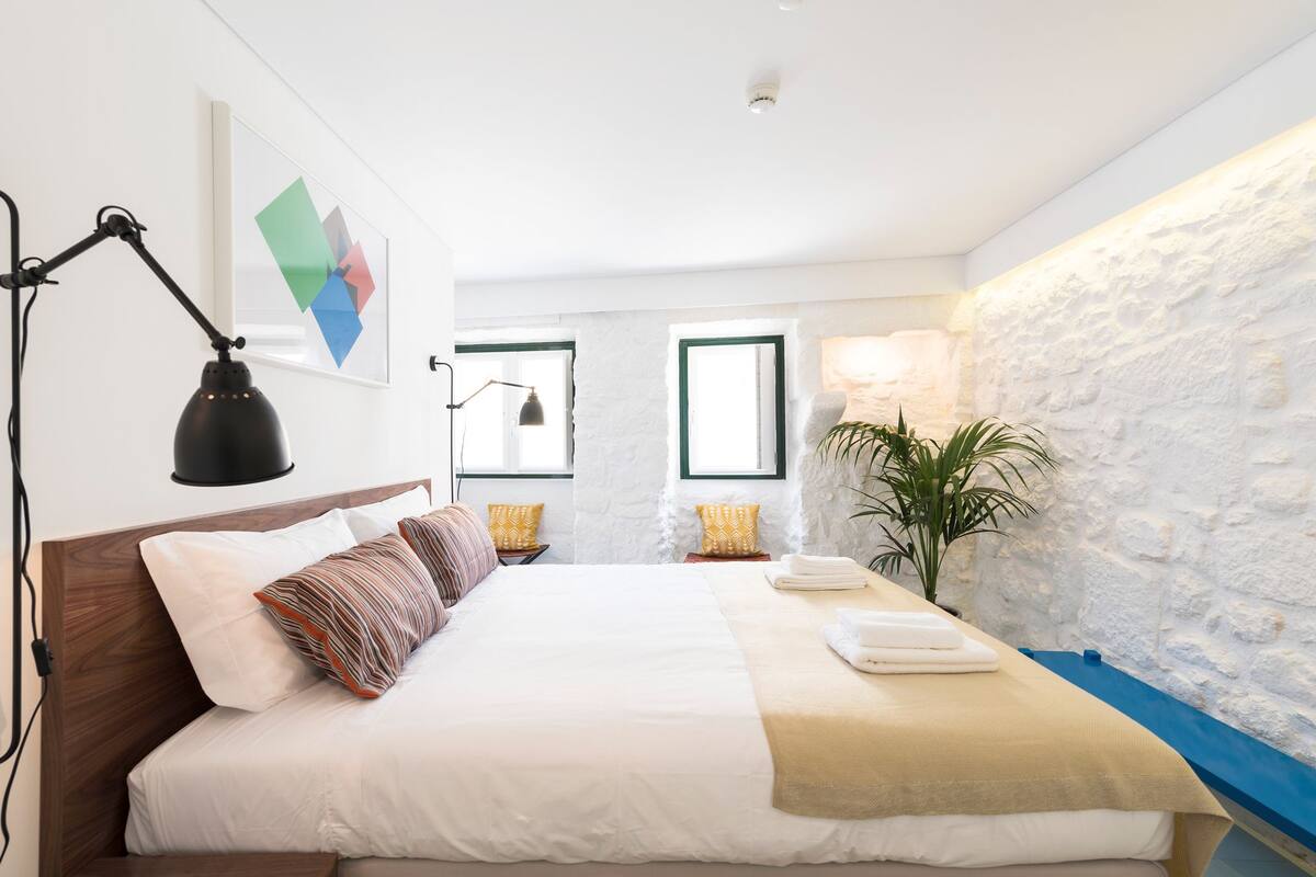 A modern bedroom with a large bed, a bedside lamp, a wall-mounted picture, and a potted plant.