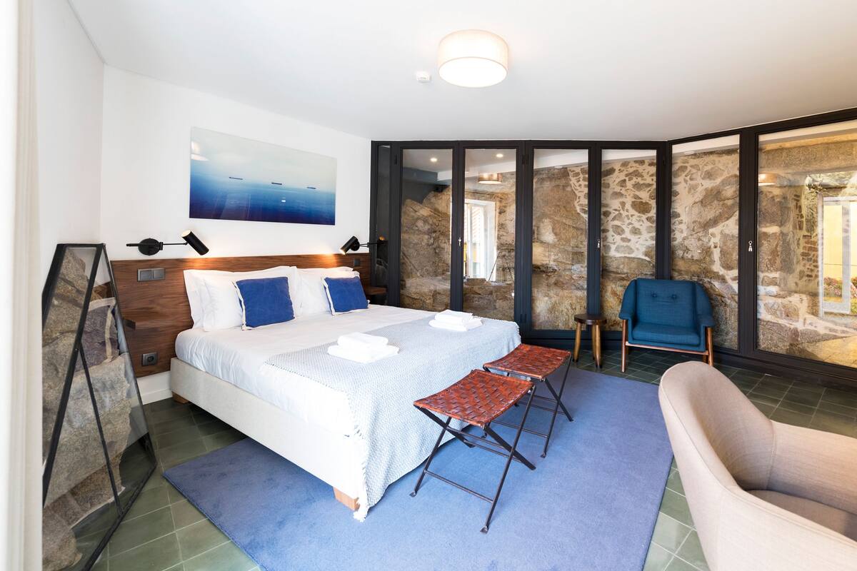 A modern bedroom with a large bed, a small table, a chair, and a view of a stone wall.