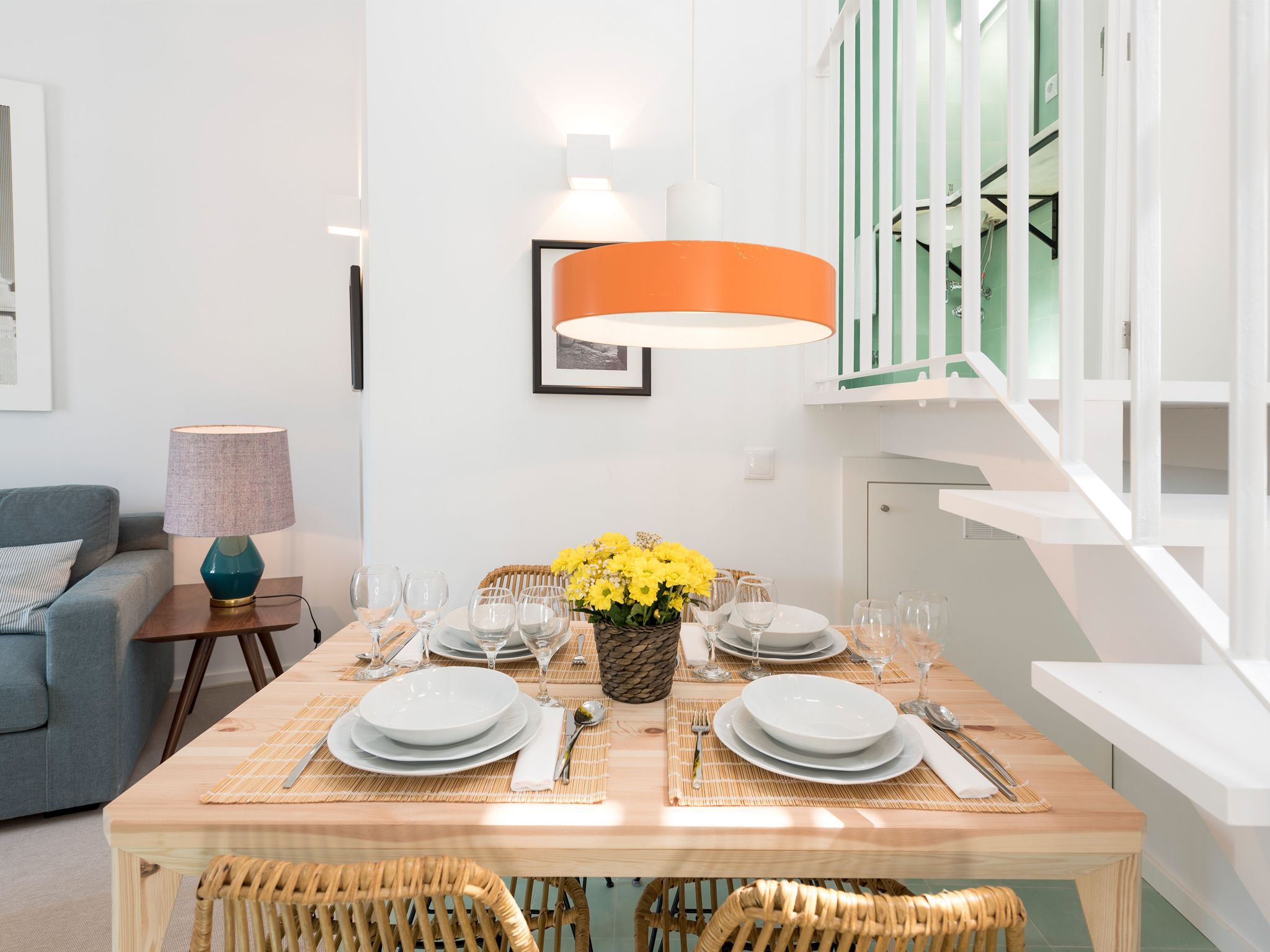 A dining area with a wooden table set for four, surrounded by wicker chairs. There is a wall-mounted orange lamp above the table, and a staircase with white railings to the right.