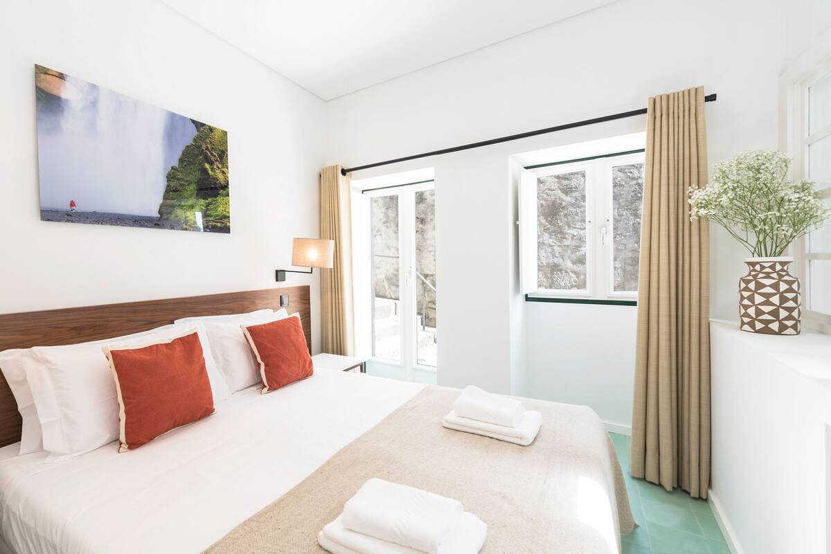A hotel room with a large bed, a view of the outdoors, a painting on the wall, and a vase with flowers.