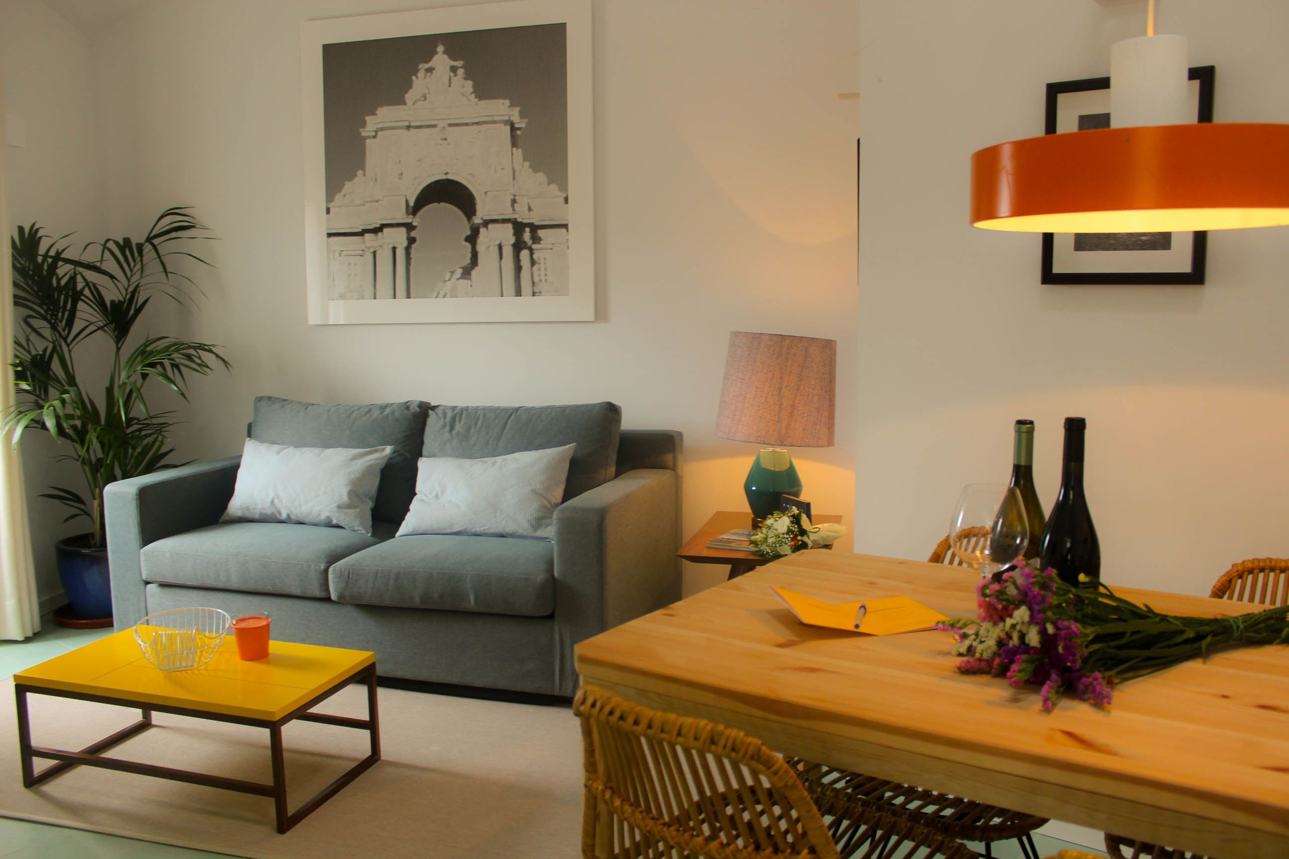 Ein Wohnzimmer mit grauem Sofa, einem gelben Couchtisch, einem Holzesstisch mit Stühlen, einer Topfpflanze und einem gerahmten Bild an der Wand.