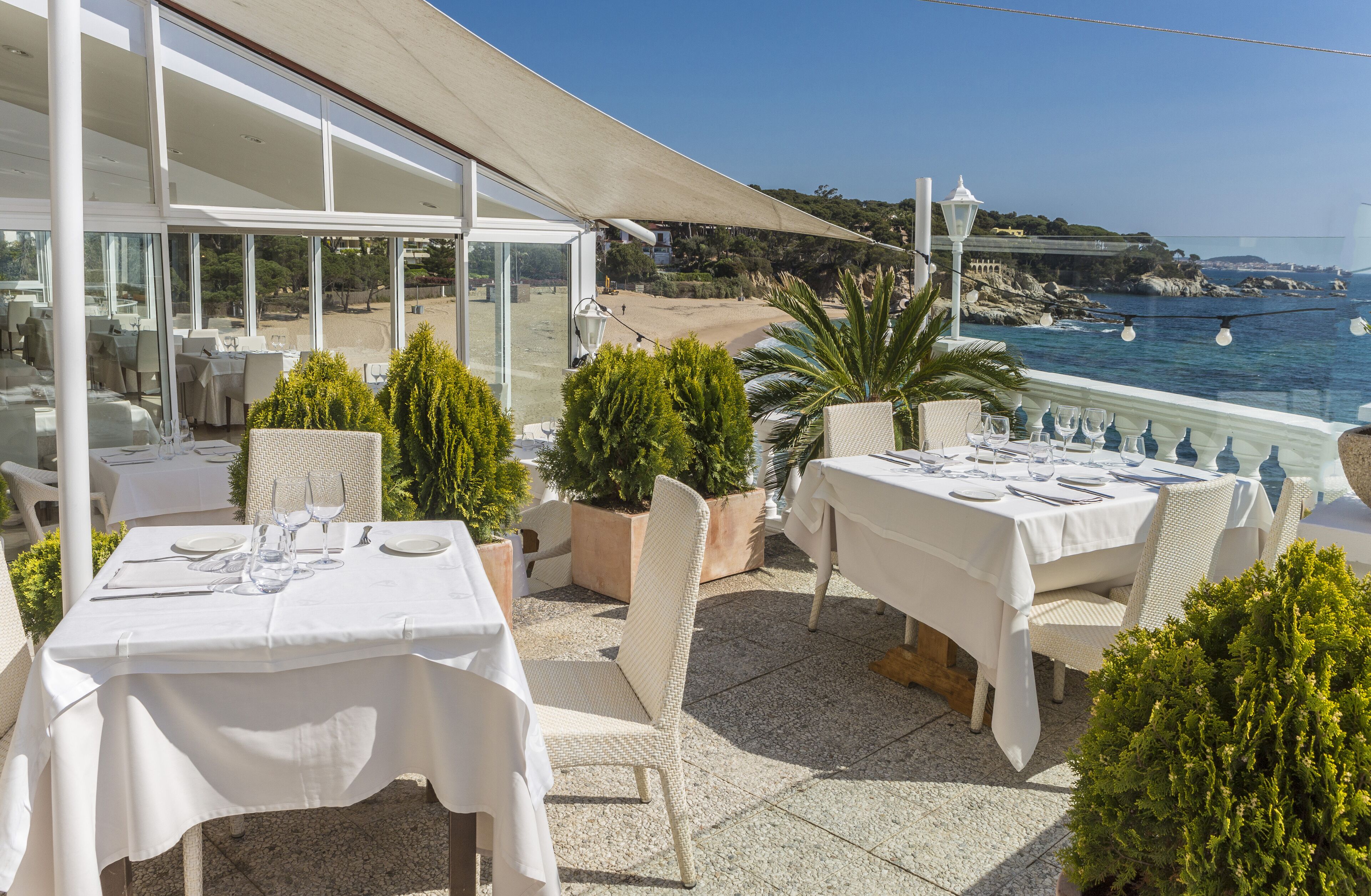 An outdoor dining area with tables set for two, overlooking a beach and the sea.