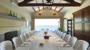 A long dining table with white chairs, set for a meal, overlooking the sea.