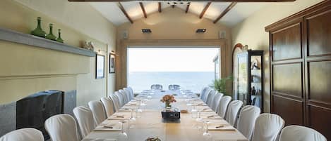 A long dining table with white chairs, set for a meal, overlooking the sea.