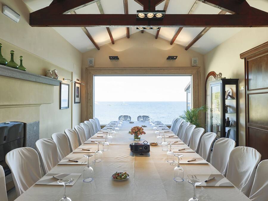 A long dining table with white chairs, set for a meal, overlooking the sea.