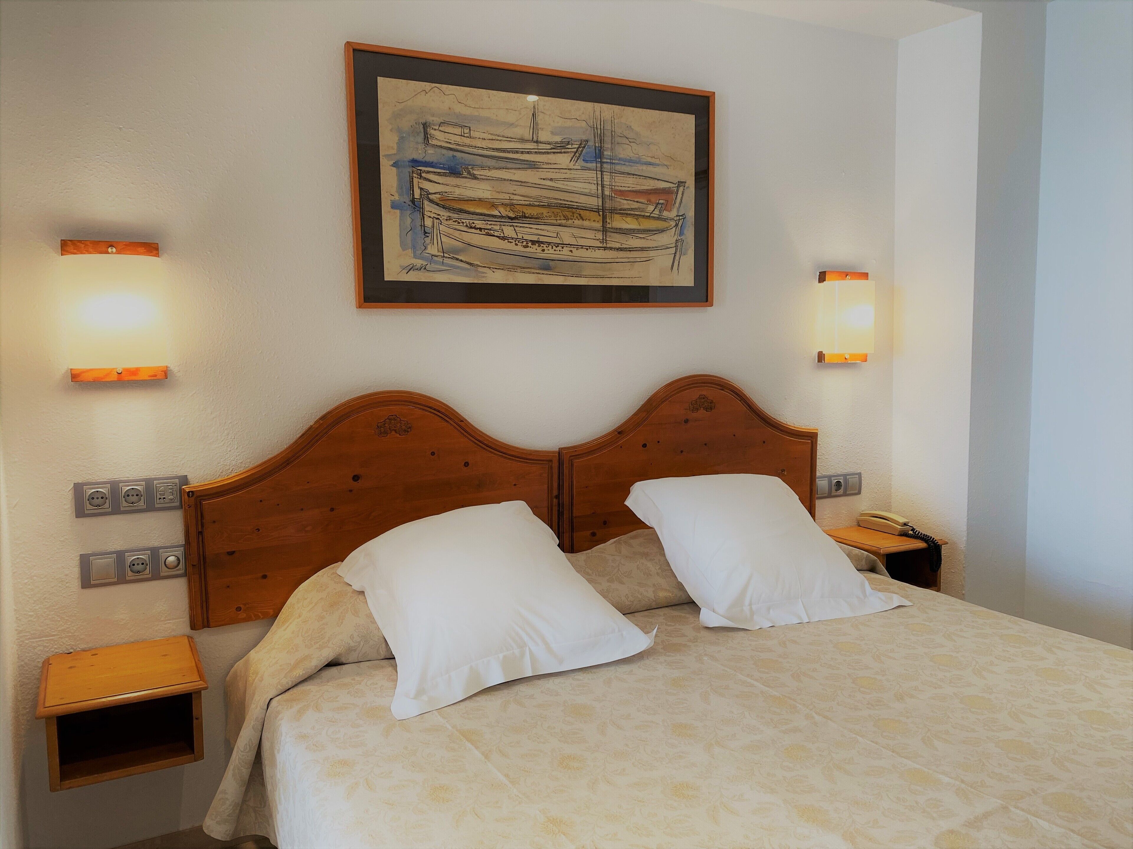 A neatly made bed with a patterned bedspread, two white pillows, and a wooden headboard. There are two bedside tables with lamps, a wall socket, and a framed artwork above the bed.