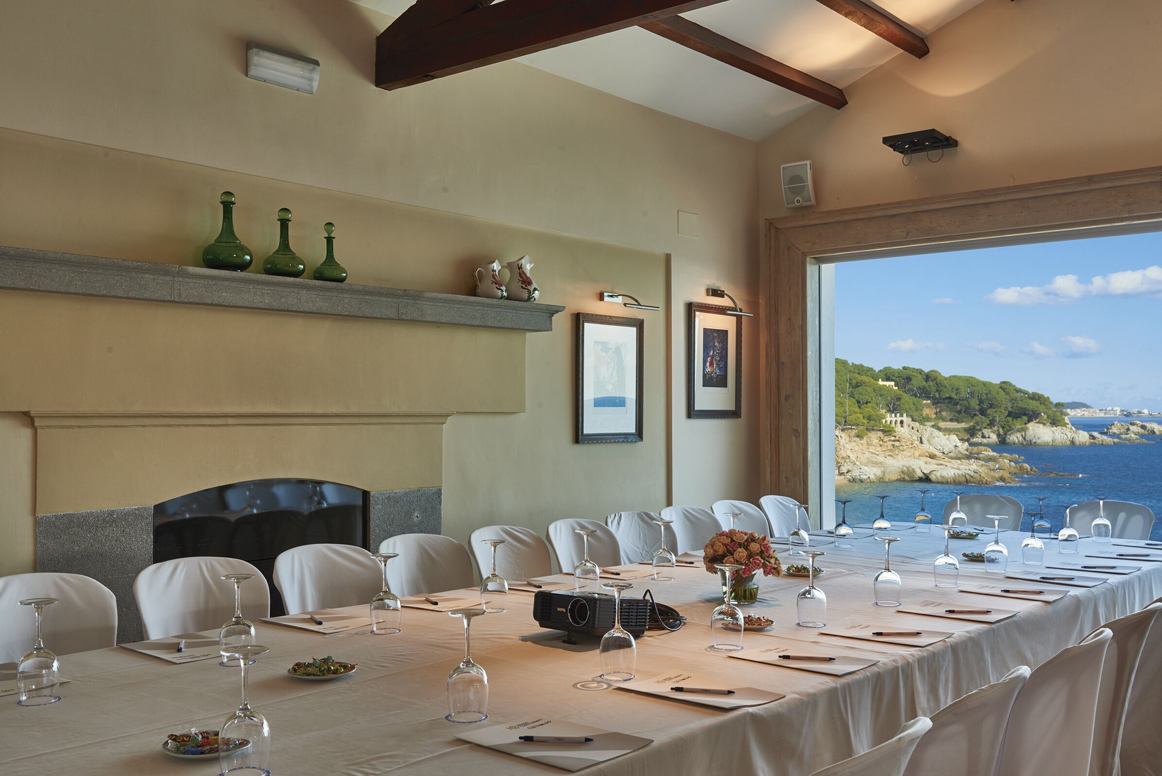 A dining area with a long table set for a meal, chairs, a fireplace, and a view of the sea.