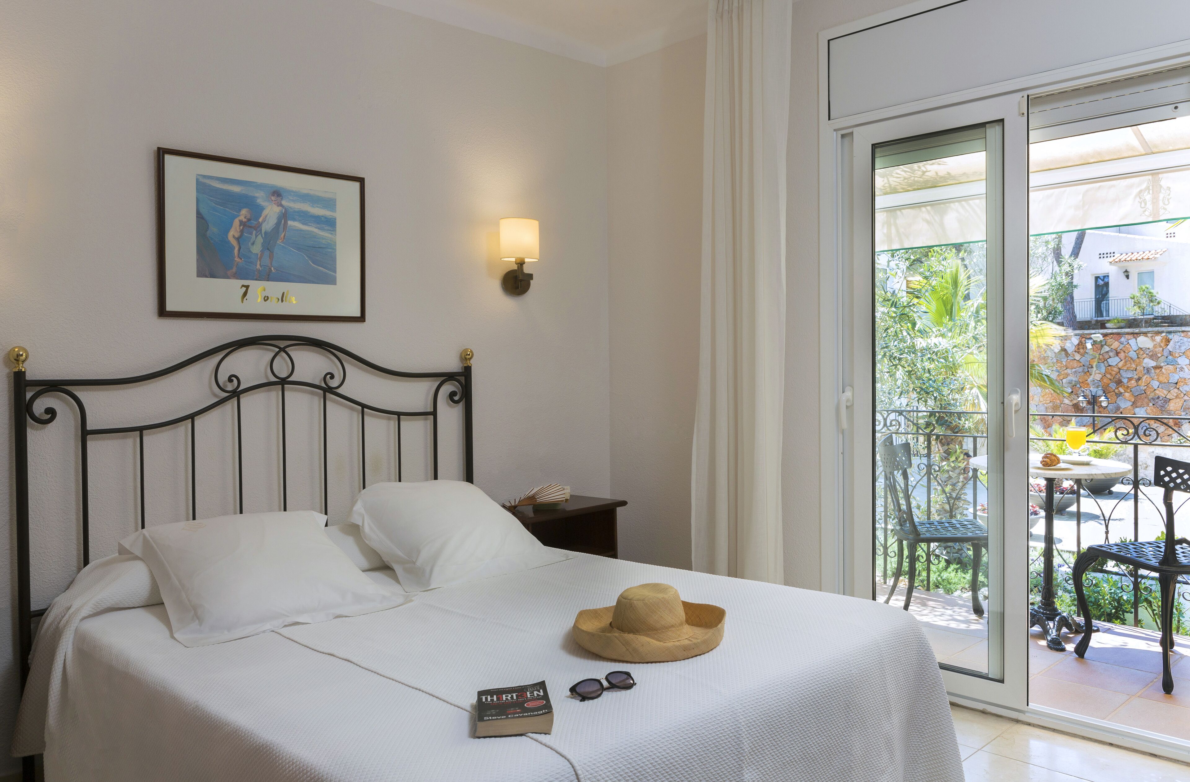 Uma cama bem arrumada com lençóis brancos, um quadro na parede, um abajur e uma varanda com móveis.