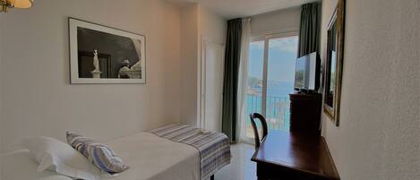 A hotel room with a bed, a desk, a chair, a TV, and a window with a view of the sea.