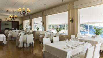 Un comedor amplio con varias mesas dispuestas para comer, ventanas grandes que ofrecen vistas al mar y candelabros elegantes.