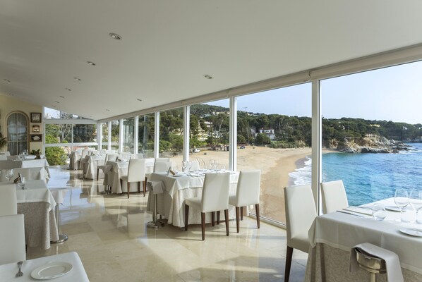 Un restaurante con mesas preparadas para comer, amplios ventanales que ofrecen vista a una playa y al océano.