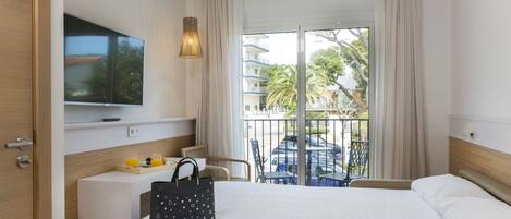 A hotel room with a bed, a TV mounted on the wall, a balcony with a view of palm trees and a car, and a desk with a chair and a handbag.