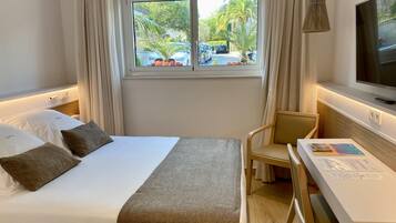 A hotel room with a bed, a desk, a chair, a TV, and a window with a view of parked cars and palm trees.