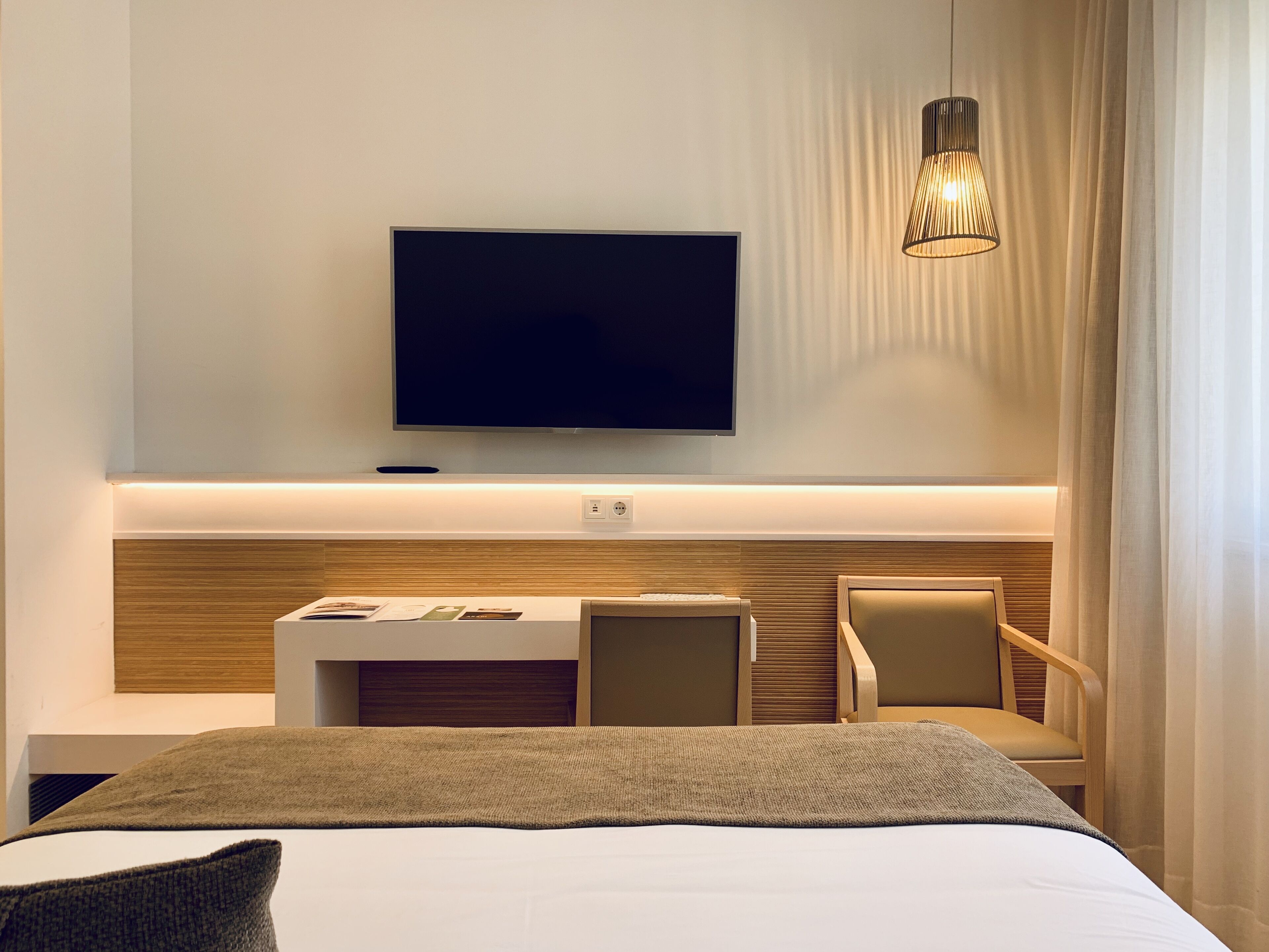 Une chambre d’hôtel moderne dotée d’un grand lit, d’un téléviseur à écran plat fixé au mur, d’un petit bureau avec une chaise et d’un luminaire suspendu.