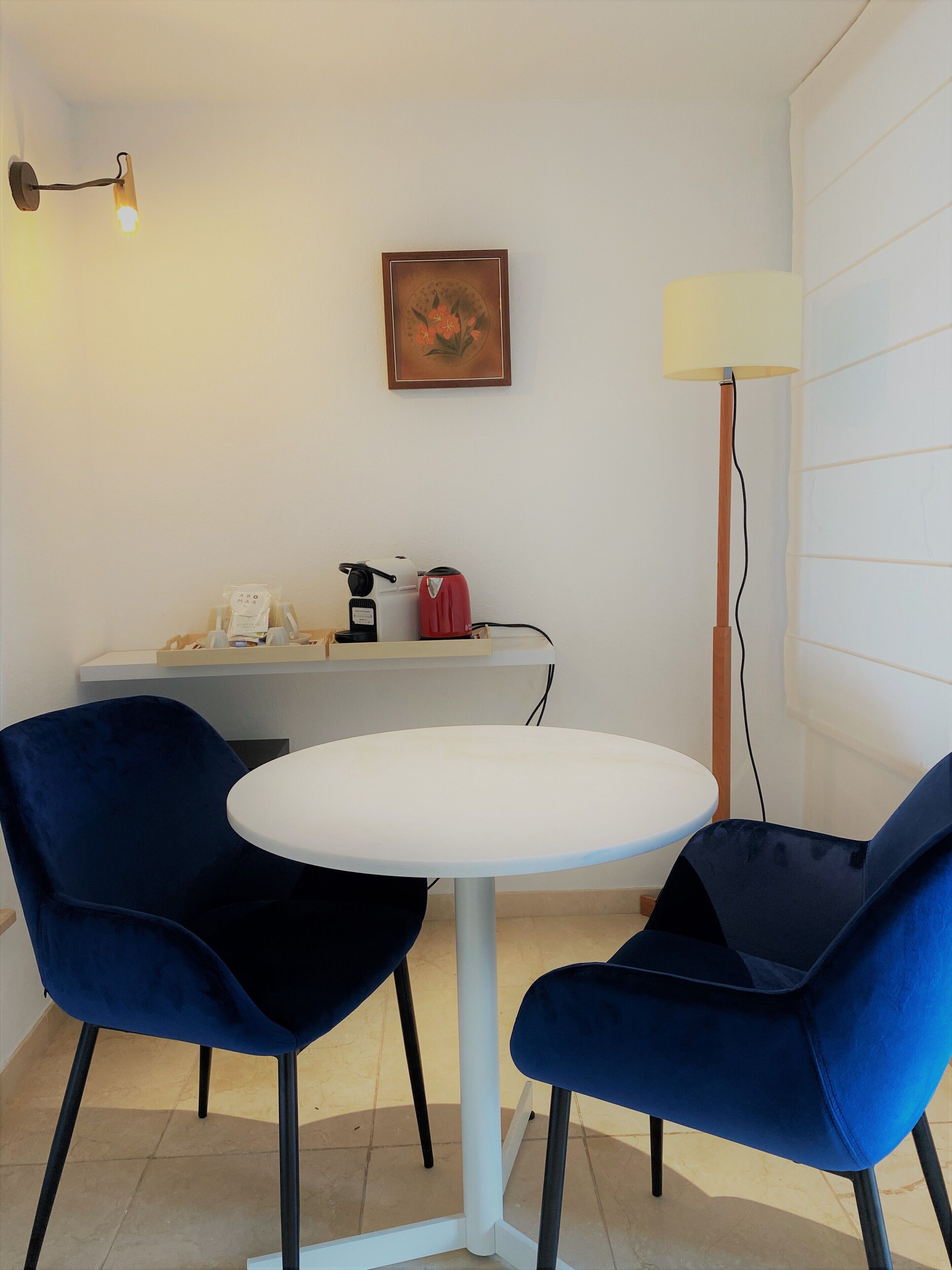 Una habitación pequeña con una mesa redonda blanca, dos sillas azules, una cafetera, un hervidor, una lámpara y un cuadro en la pared.