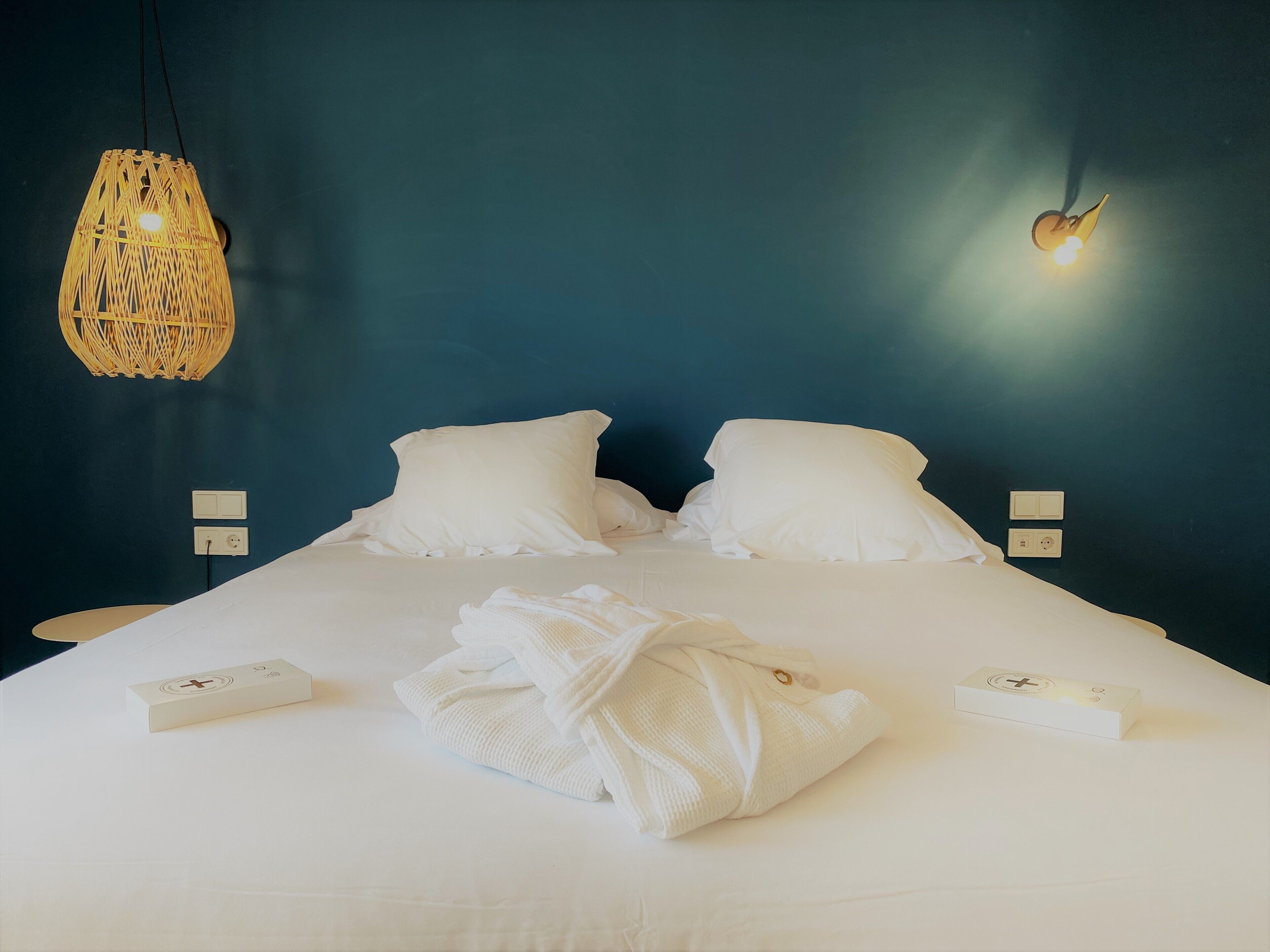 A neatly made bed with white linens, a woven light fixture above it, and two wall-mounted light switches.