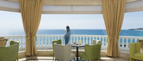 A balcony with a view of the sea, furnished with green chairs and a table, and a person standing by the railing.