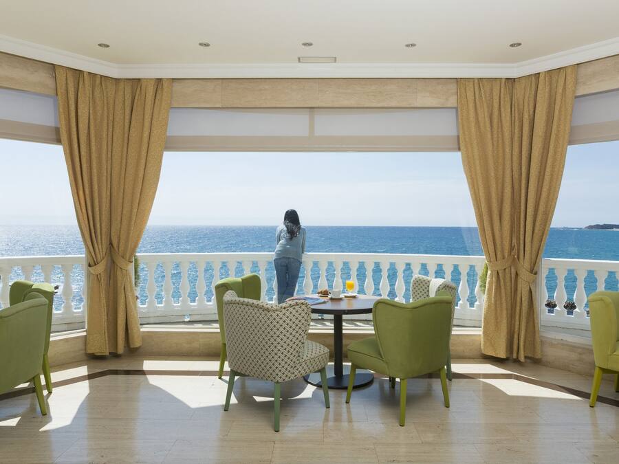 Un balcon donnant sur la mer, agrémenté de chaises vertes et d’une table, et une personne se tenant près de la balustrade.