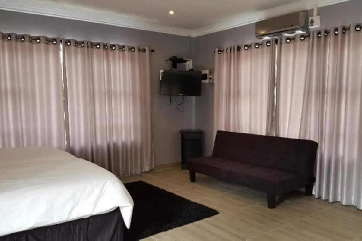 A hotel room with a bed, a sofa, a flat-screen TV mounted on the wall, and curtains.